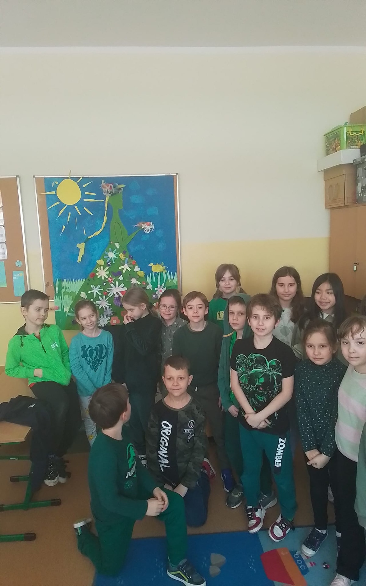 Uczniowie w klasie pozują do zdjęcia, każdy ma zielony element ubioru z okazji pierwszego dnia wiosny. W tle plakat z błękitnym niebem i słońcem i Panią Wiosną.