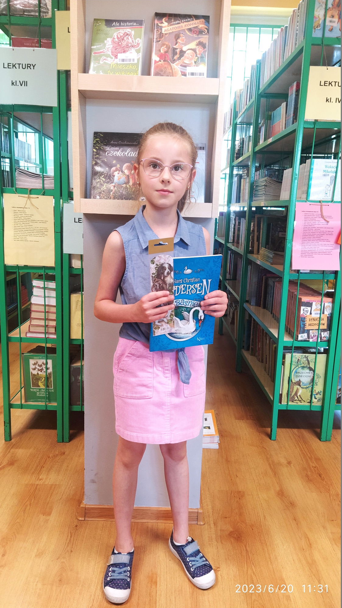 Dziewczynka stoi w bibliotece przy regałach z książkami. W ręce prezentuje nagrodę książkową, którą otrzymała za czytelnictwo.