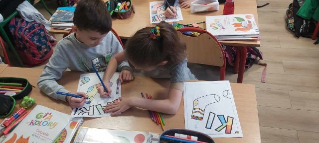Dwójka dzieci chłopiec i dziewczynka siedzą przy ławce, kolorują obrazek ze skarpetką i literkami.