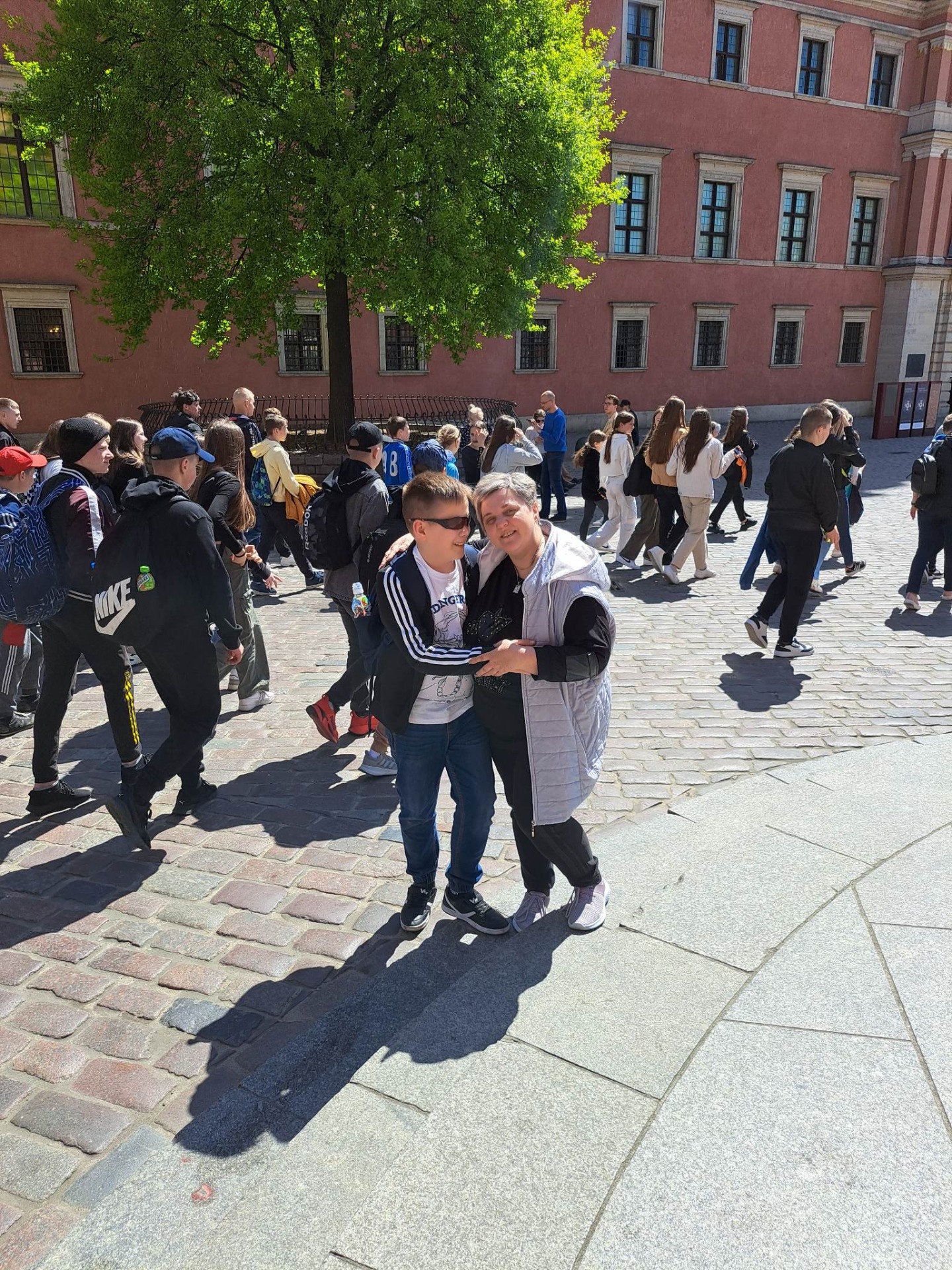 Wycieczka uczniów idzie na tle Zamku Królewskiego w Warszawie. Na pierwszym planie stoi uczeń z opiekunką. Trzymają się  za ręce i uśmiechają do zdjęcia
