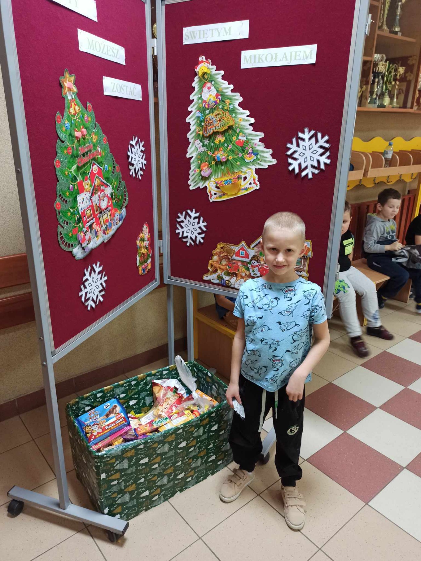 Dzieci wrzucają słodkie upominki do pudełka z darami obok gazetki z napisem "I Ty możesz zostać Świętym Mikołajem".