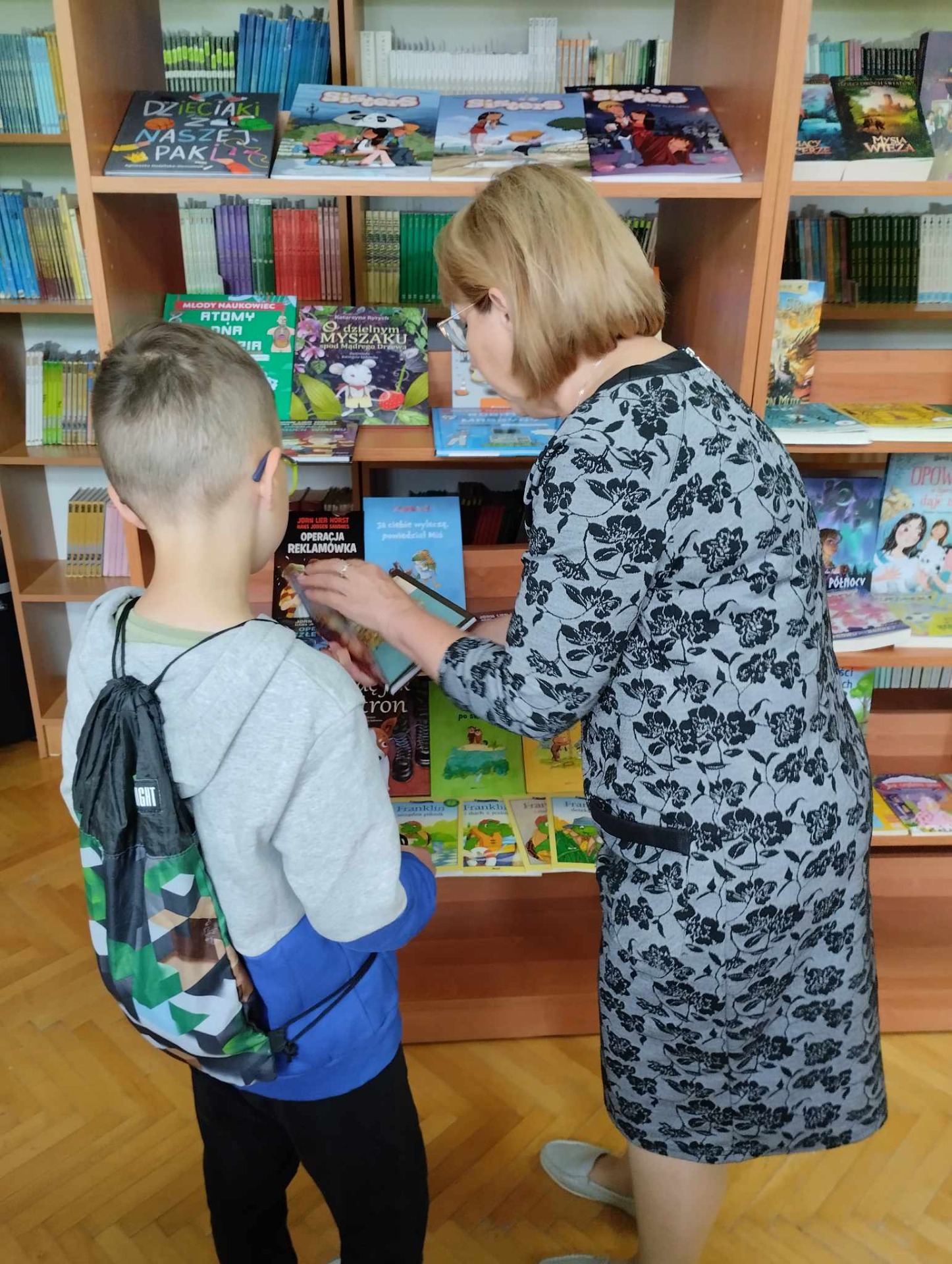 Fotografia przestawia bibliotekę szkolną, a w niej ucznia, który wraz z panią bibliotekarką stoją przed regałem z nowościami bibliotecznymi.