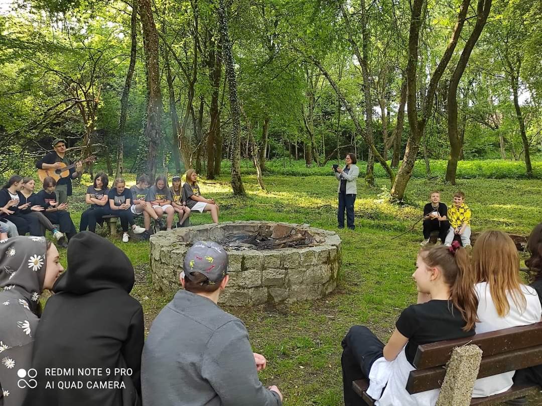 Na zdjęciu osiemnastu uczniów siedzi wokół ogniska. Mężczyzna gra na gitarze, a kobieta robi zdjęcie. Wokół rosną drzewa.