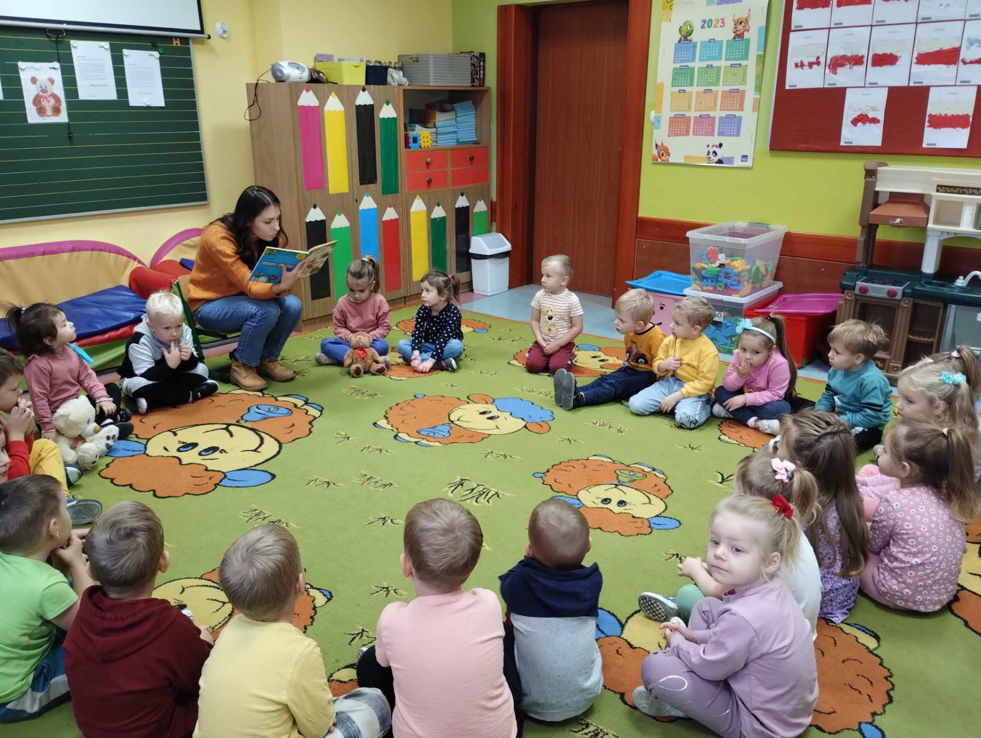 Przedszkolaki siedzą na kolorowym dywanie i słuchają książki czytanej przez p. Olgę.