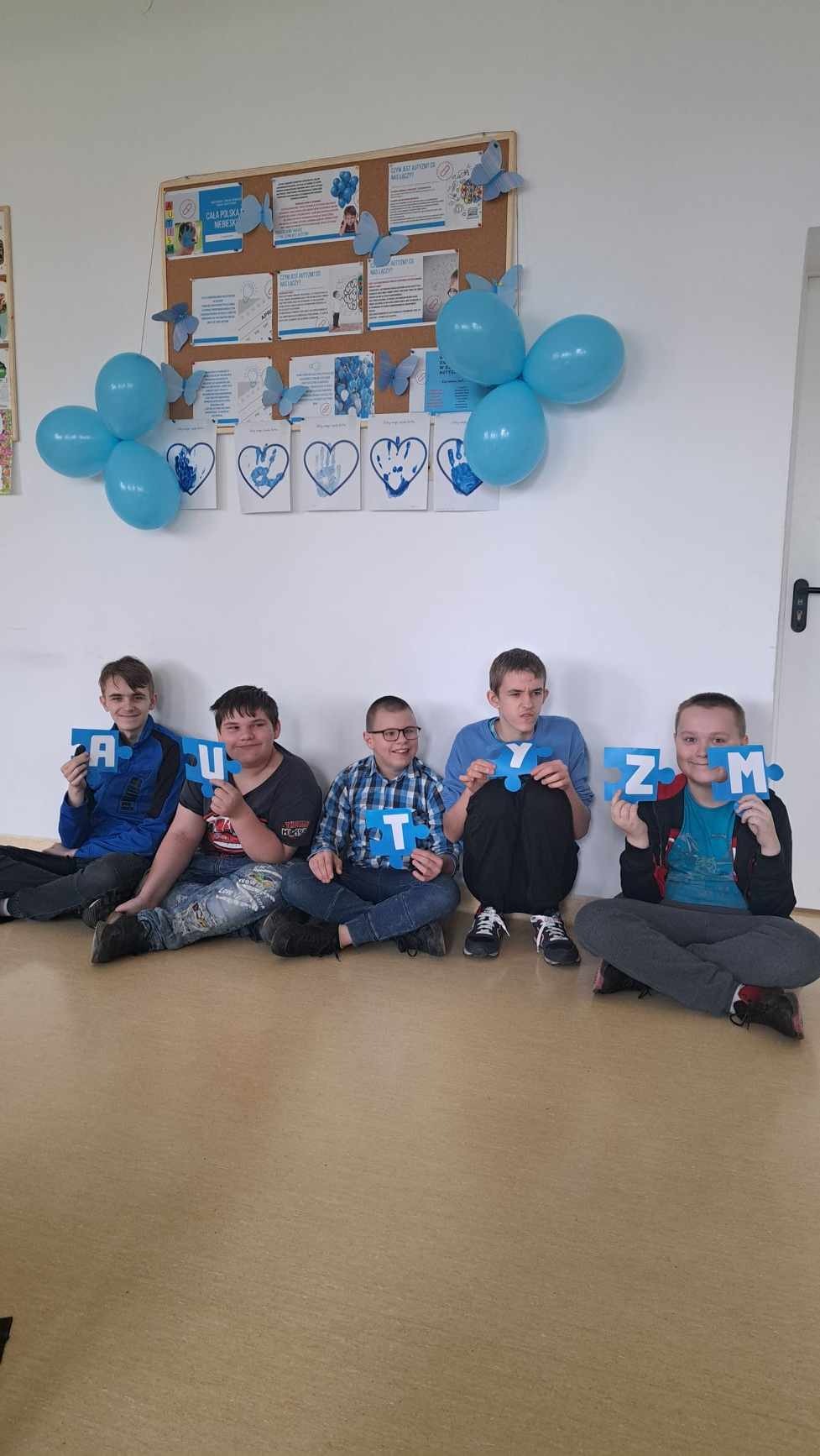 5 uczniów ubranych w elemnty koloru niebieskiego siedzi "po turecku" pod ścianą , w ręku trzymają litery składające się na słowo autyzm - nad ich glowmi wiszą niebieskie balony