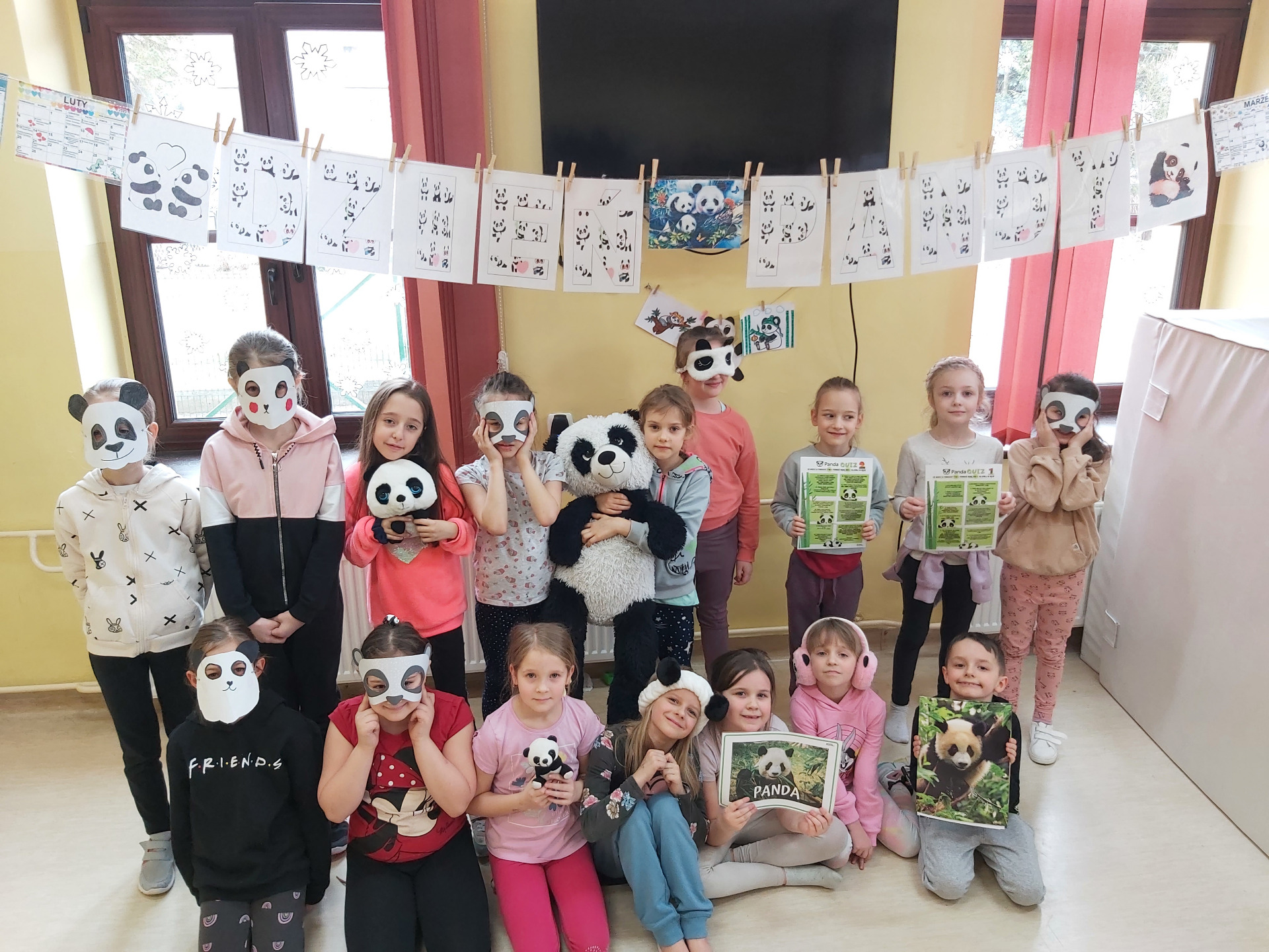 Zdjęcie przedstawia dzieci stojące pod napisem Dzień Pandy. Niektóre z dzieci trzymają w rękach maskotki pandy, inne zdjęcia przedstawiające wizerunek pandy wielkiej, jeszcze inne mają na twarzach pandzie maski.