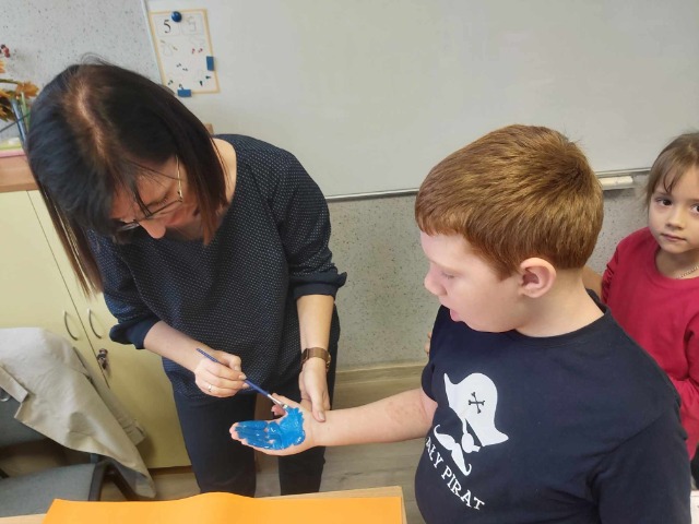 Nauczycielka maluje chłopcu dłoń niebieską farbą, obok chłopca stoi dziewczynka.