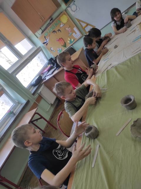 6 uczniów siedzi przy stole zakrytym obrusem i lepi swoje kubki z gliny