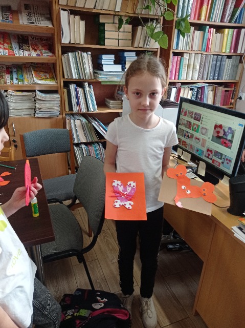 W bibliotece szkolnej stoi dziewczynka, w rękach trzyma kartki z wyklejonymi obrazkami.
