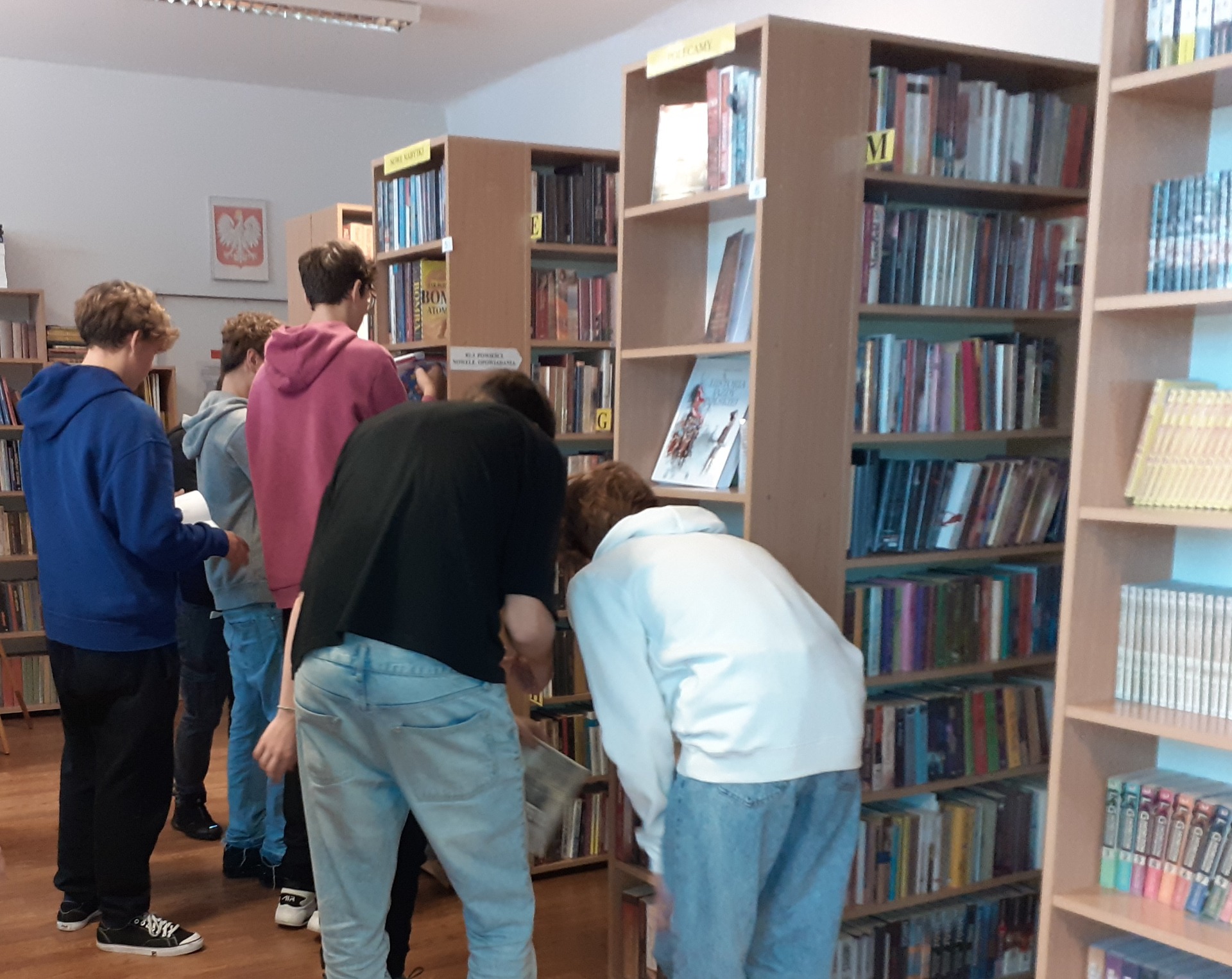 Grupa uczniów poszukuje książek na regałach bibliotecznych.