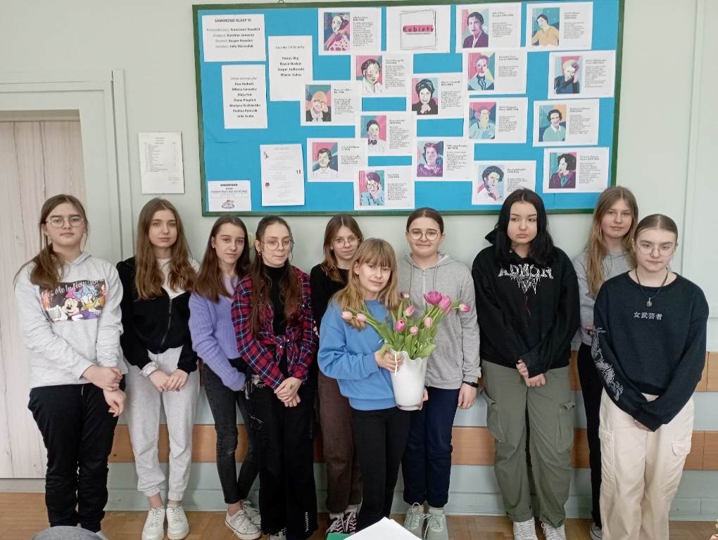 Na zdjęciu szóstym stoi dziesięć uczennic klasy szóstej. Jedna z nich trzyma wazon z kwiatami. Za nimi na ścianie wisi gazetka klasowa przedstawiająca "Kobiety, które zmieniły świat".