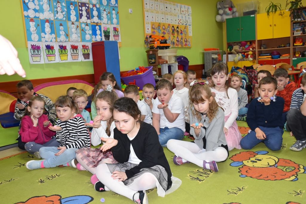 Grupa dzieci siedzi na kolorowym dywanie i wykonuje ćwiczenia rozluźniające narządy głosowe.