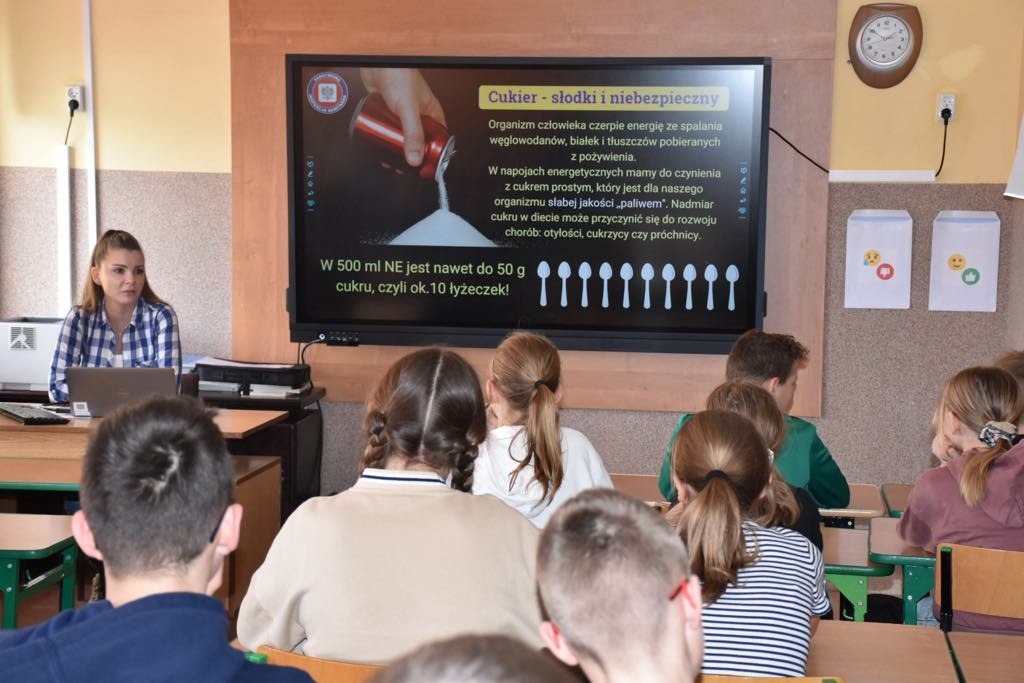 Uczniowie na spotkaniu z gośćmi z SANEPID-u oglądają prezentację multimedialną wyświetlaną na monitorze interaktywnym.