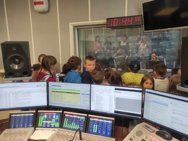 Sprzęt radiowy za nim grupa dzieci wpatrującą się w okno nad którym wisi elektroniczny zegar, za którym siedzi Pani w słuchawkach, za ekranem komputera, a przed nią znajduje się mikrofon.
