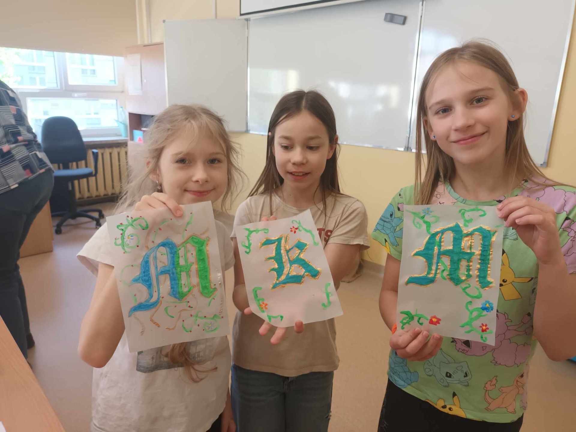 Uczennice prezentują swoje prace z warsztatów. Trzy dziewczynki trzymają w rękach kartki na których różnymi kolorami są narysowane ozdobne litery. Dziewczyny stoją w klasie szkolnej, w tle widać fotel nauczyciela i tablicę multimedialną.