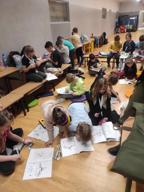 grupa dzieci siedzi przy stolikach lub na podłodze, rozwiązując książeczki