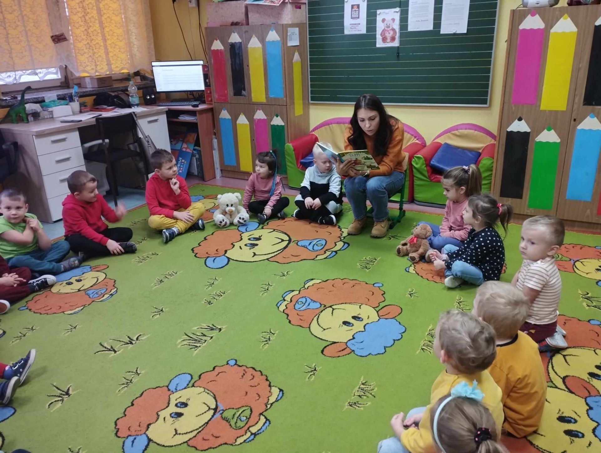 Przedszkolaki siedzą na kolorowym dywanie i słuchają książki czytanej przez p. Olgę.