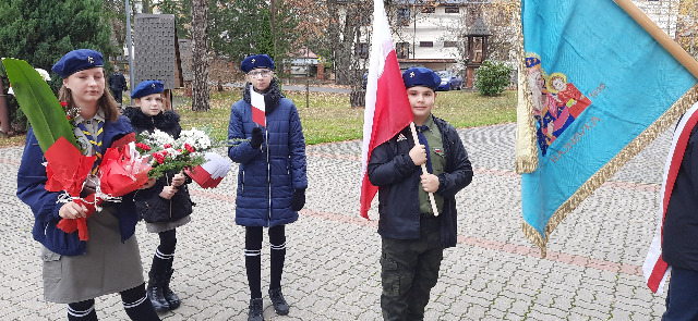 Stoi czworo dzieci ubranych w mundurki harcerskie, trzy dziewczynki i jeden chłopiec. Dzieci trzymają w ręku flagi Polski.