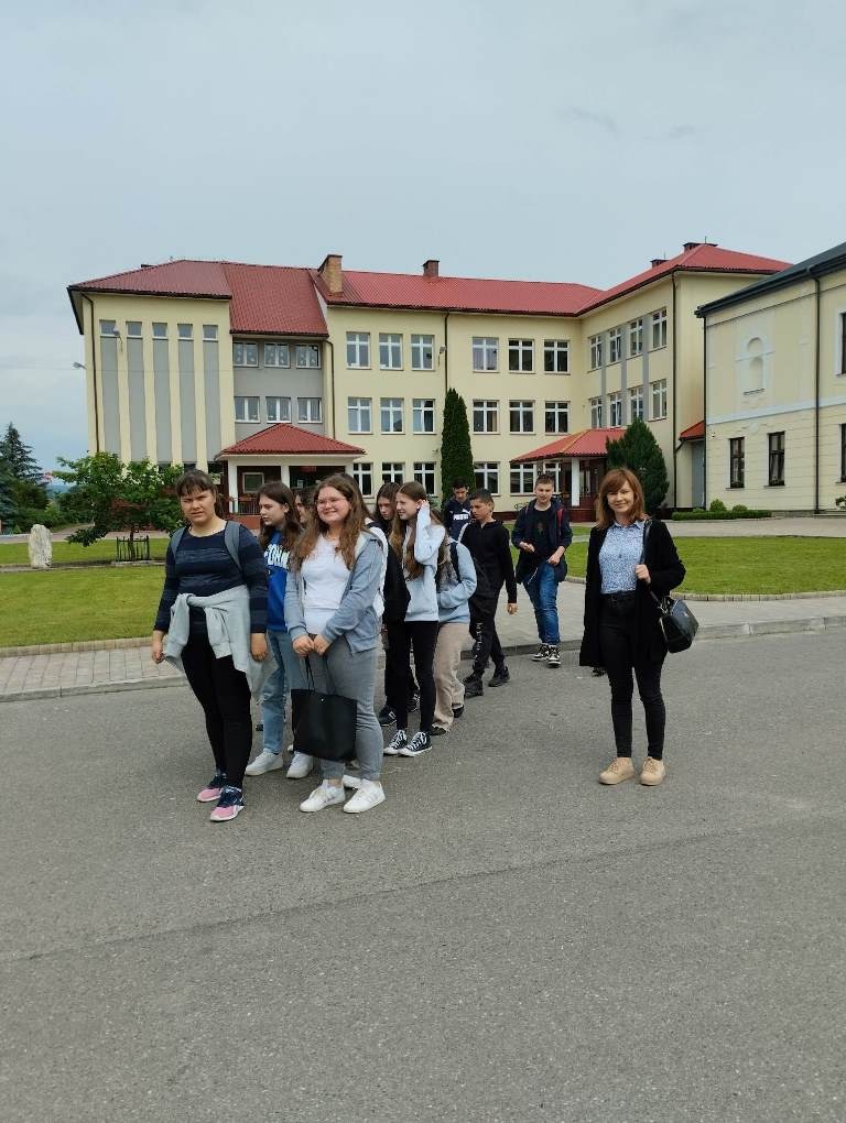 Zdjęcie przedstawia piętnastu uczniów klasy siódmej, którzy wraz z wychowawczynią idą na wycieczkę. W tle Budynek szkoły.